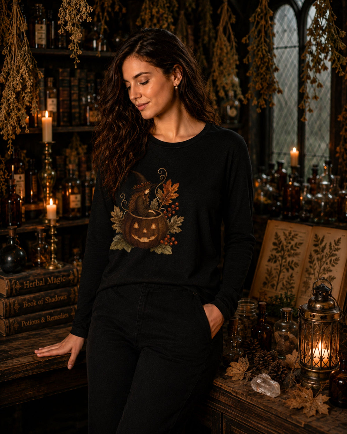 A woman wearing a black long-sleeve crewneck sweatshirt with a pumpkin graphic, standing in a dark, atmospheric room decorated with candles, books, and autumn foliage.