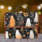 A structured handbag with a black background and a whimsical print of orange and white ghosts, white flowers, and small birds, accented with brown leather trim and a top handle, set against a warm, blurred bokeh background on a wooden surface.