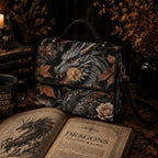 A structured handbag with a dark fantasy dragon illustration, displayed on a rustic table with an open book, a mug, and dried flowers in the background.
