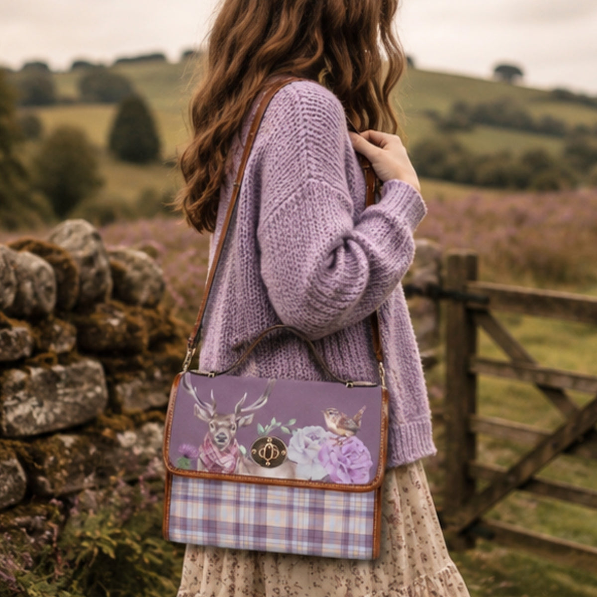 A woman in a lavender knit sweater and floral skirt carries a lavender crossbody bag with a deer and floral print on the front and a purple plaid lower section, set against a blurred outdoor landscape of rolling green hills and a stone wall.