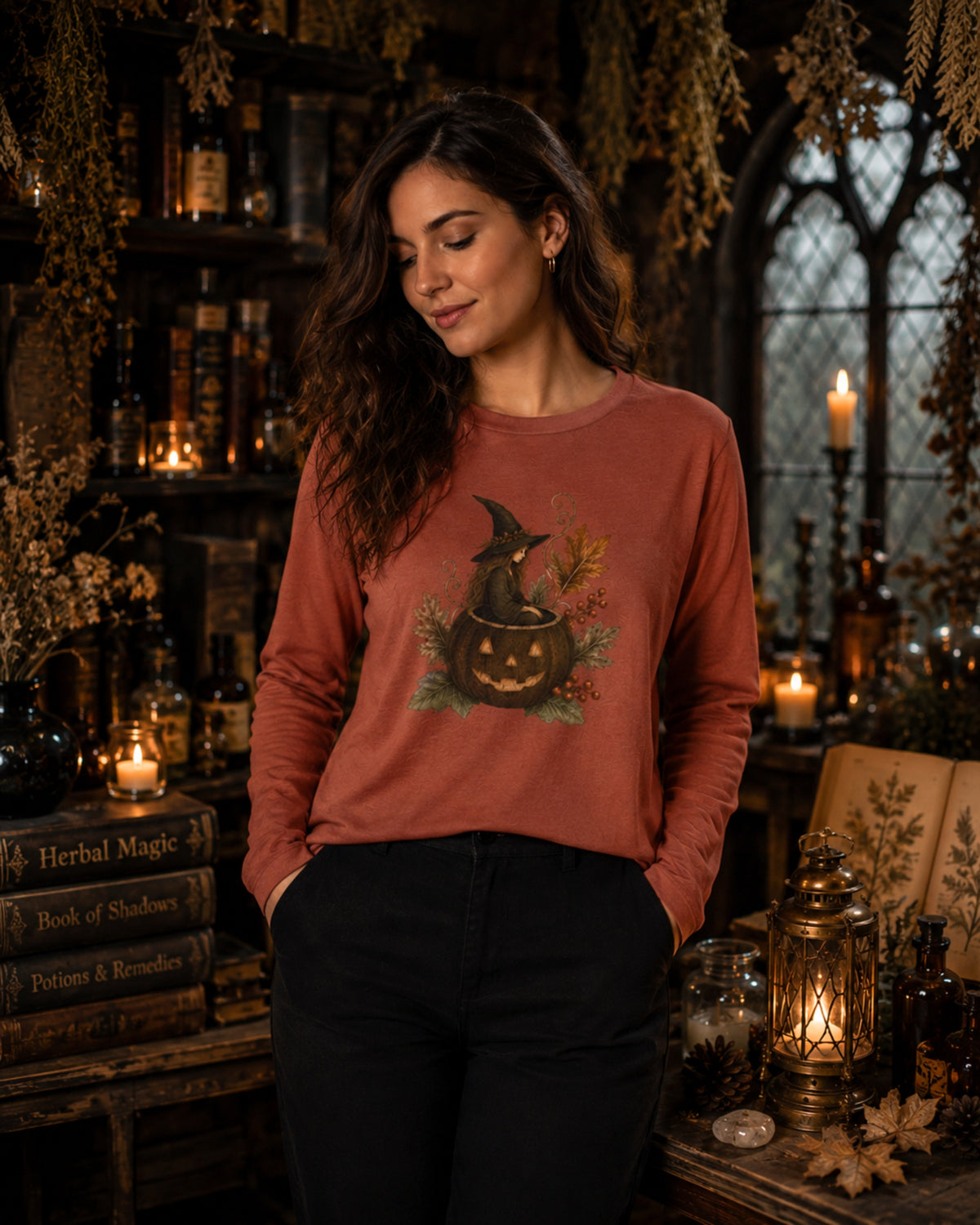 A woman wearing a rust-orange long-sleeve t-shirt with a Halloween graphic of a witch sitting on a jack-o'-lantern, posed in a cozy, dimly lit library or study filled with books, candles, and lanterns.