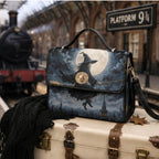 A witch-themed handbag with a printed illustration of a witch riding a broomstick under a full moon, styled on top of a vintage suitcase with a black scarf in the foreground and a blurred train station platform background.