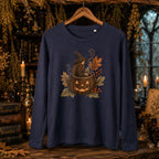 A navy blue long-sleeve t-shirt hanging on a wooden hanger against a dark, atmospheric Halloween-themed background with candles, books, and dried foliage. The shirt features a central graphic of a witch sitting in a jack-o'-lantern pumpkin surrounded by autumn leaves and berries.