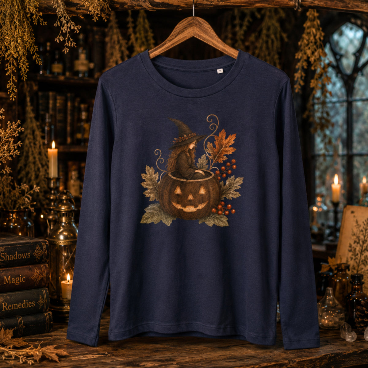 A navy blue long-sleeve t-shirt hanging on a wooden hanger against a dark, atmospheric Halloween-themed background with candles, books, and dried foliage. The shirt features a central graphic of a witch sitting in a jack-o'-lantern pumpkin surrounded by autumn leaves and berries.