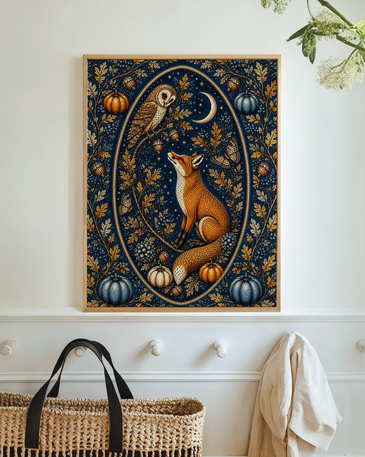 A framed vertical art print featuring a fox, owl, moon, and pumpkins in an ornate navy and gold pattern, displayed on a white wall above a woven basket.