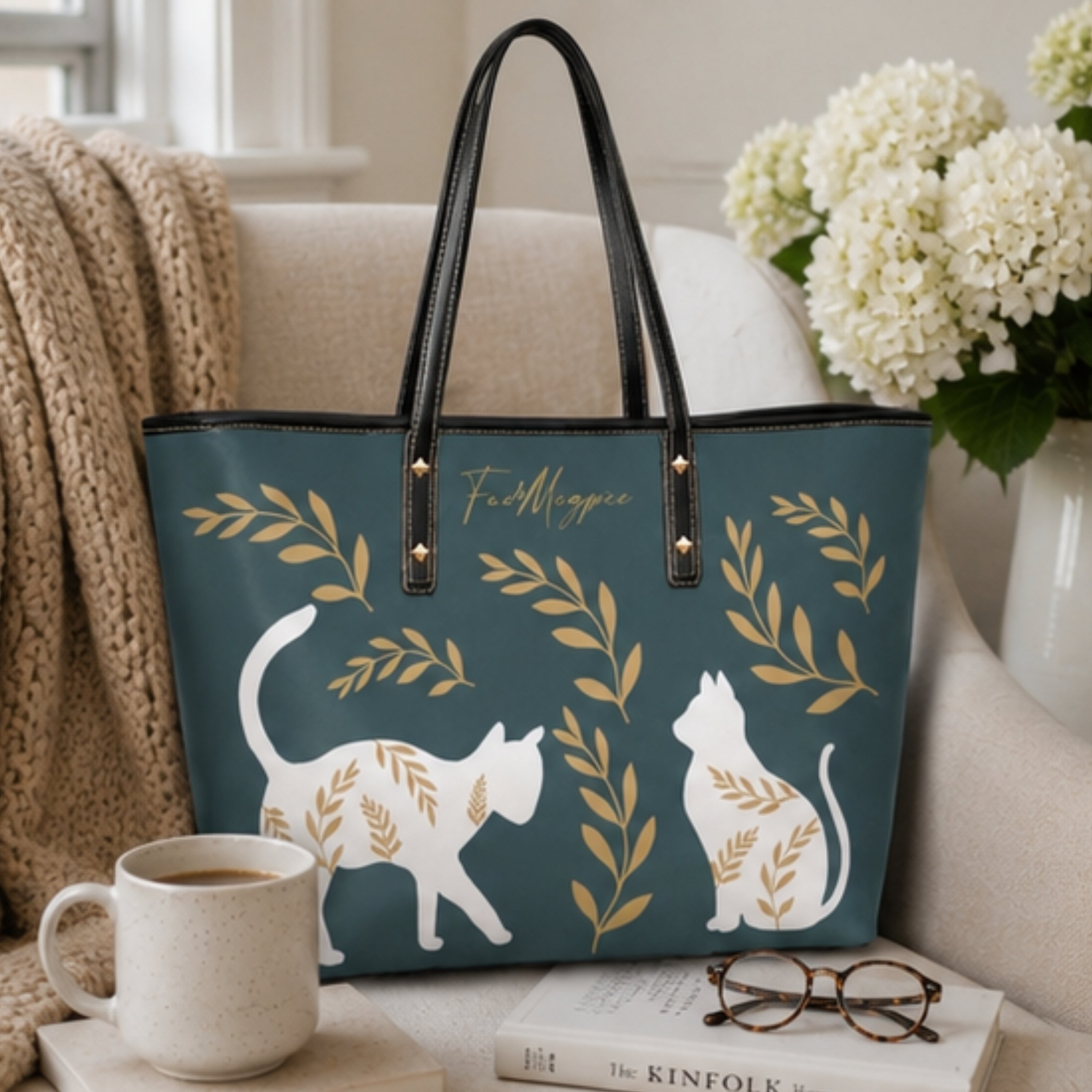 A teal tote bag with black handles and gold botanical motifs, featuring white cat silhouettes and the script text 'Fa Mignonne'. The bag is styled on a sofa next to a mug, glasses, a book, and white flowers.