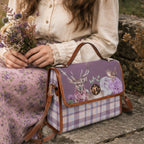A purple and lavender plaid handbag with a deer and floral print on the front flap, styled on a stone surface next to a person wearing a cream blouse and a purple floral skirt.
