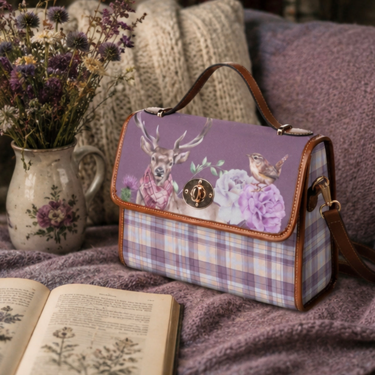 A purple and lavender plaid handbag with a flap cover featuring a deer, flowers, and a bird print, styled on a cozy blanket next to a vase of dried flowers and an open book.