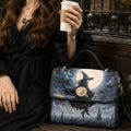 A woman in a black dress holds a white coffee cup beside a structured handbag printed with a witch riding a broomstick in front of a full moon and castle skyline.