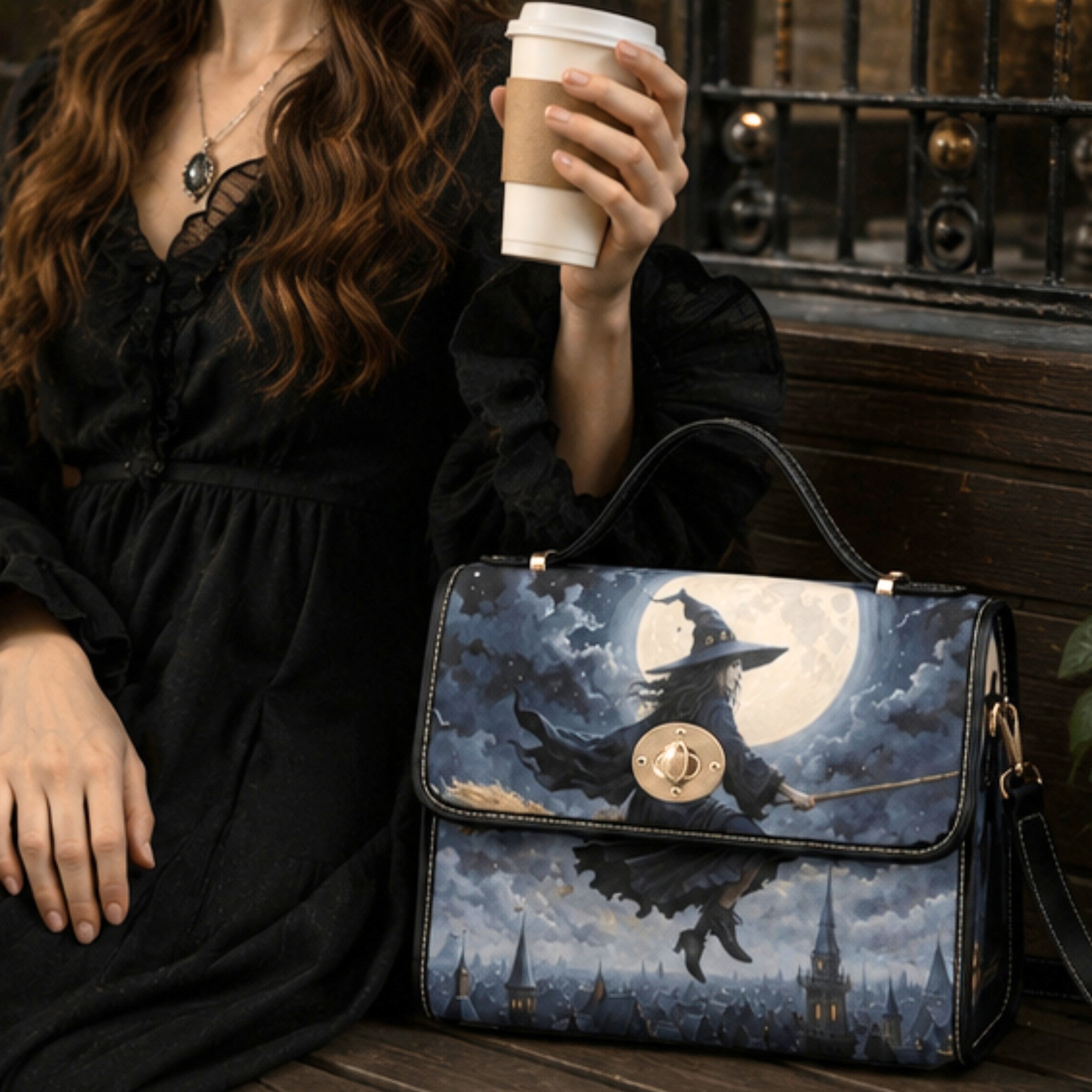 A woman in a black dress holds a white coffee cup beside a structured handbag printed with a witch riding a broomstick in front of a full moon and castle skyline.