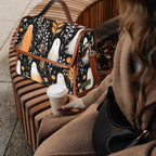 A woman in a brown coat sits on a curved wooden bench holding a takeaway coffee cup, with a black patterned shoulder bag resting on the bench beside her. The bag features a whimsical print of white ghosts, orange birds, and white birds on a black background with brown leather trim.