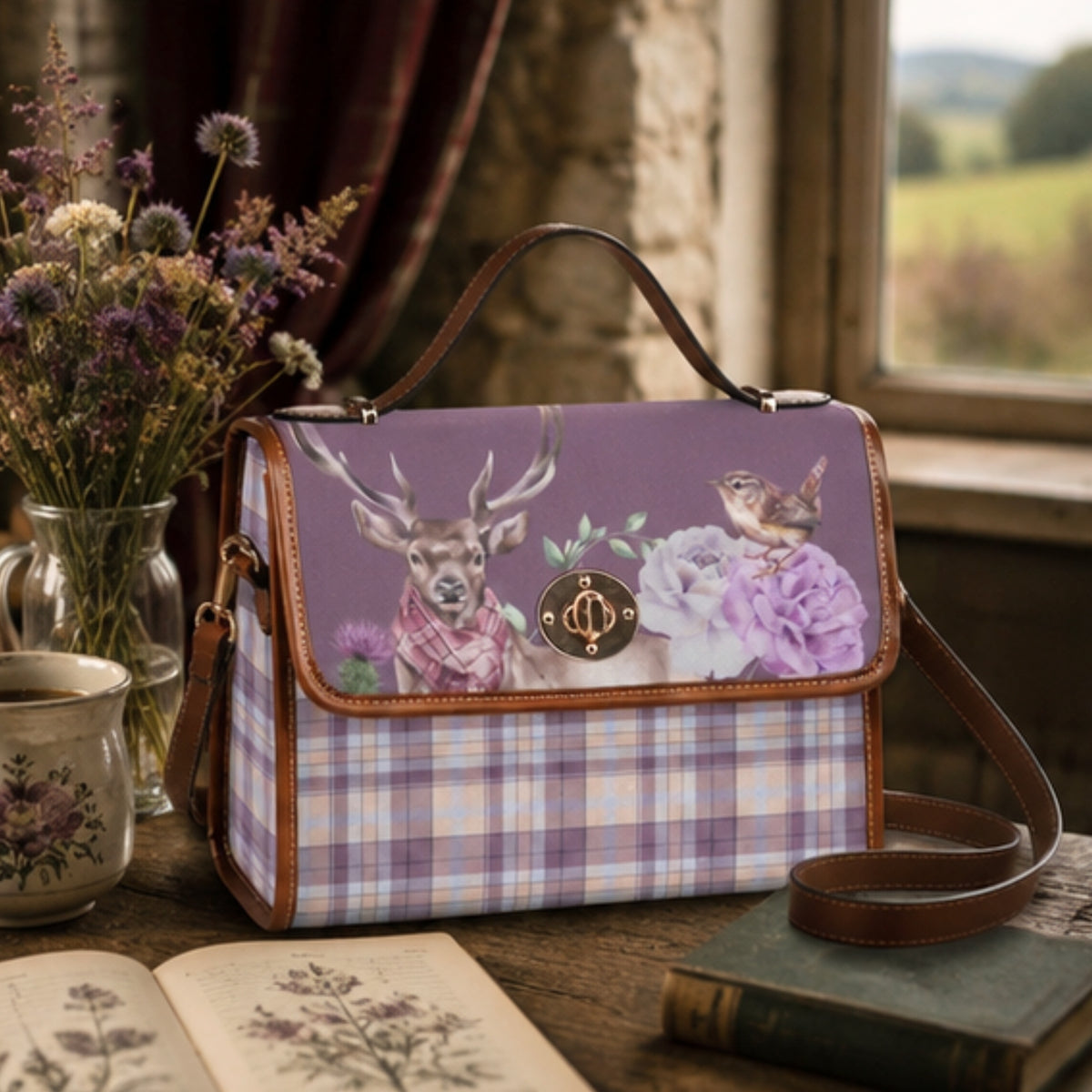 A purple and lavender plaid handbag with a floral and deer print on the front flap, styled on a rustic wooden table with dried flowers, an open book, and a mug in a cozy indoor setting.