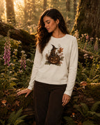 A woman stands in a sunlit forest wearing a cream long-sleeve crewneck sweatshirt with a Halloween-themed graphic of a witch's cauldron and jack-o'-lantern, paired with dark brown pants.