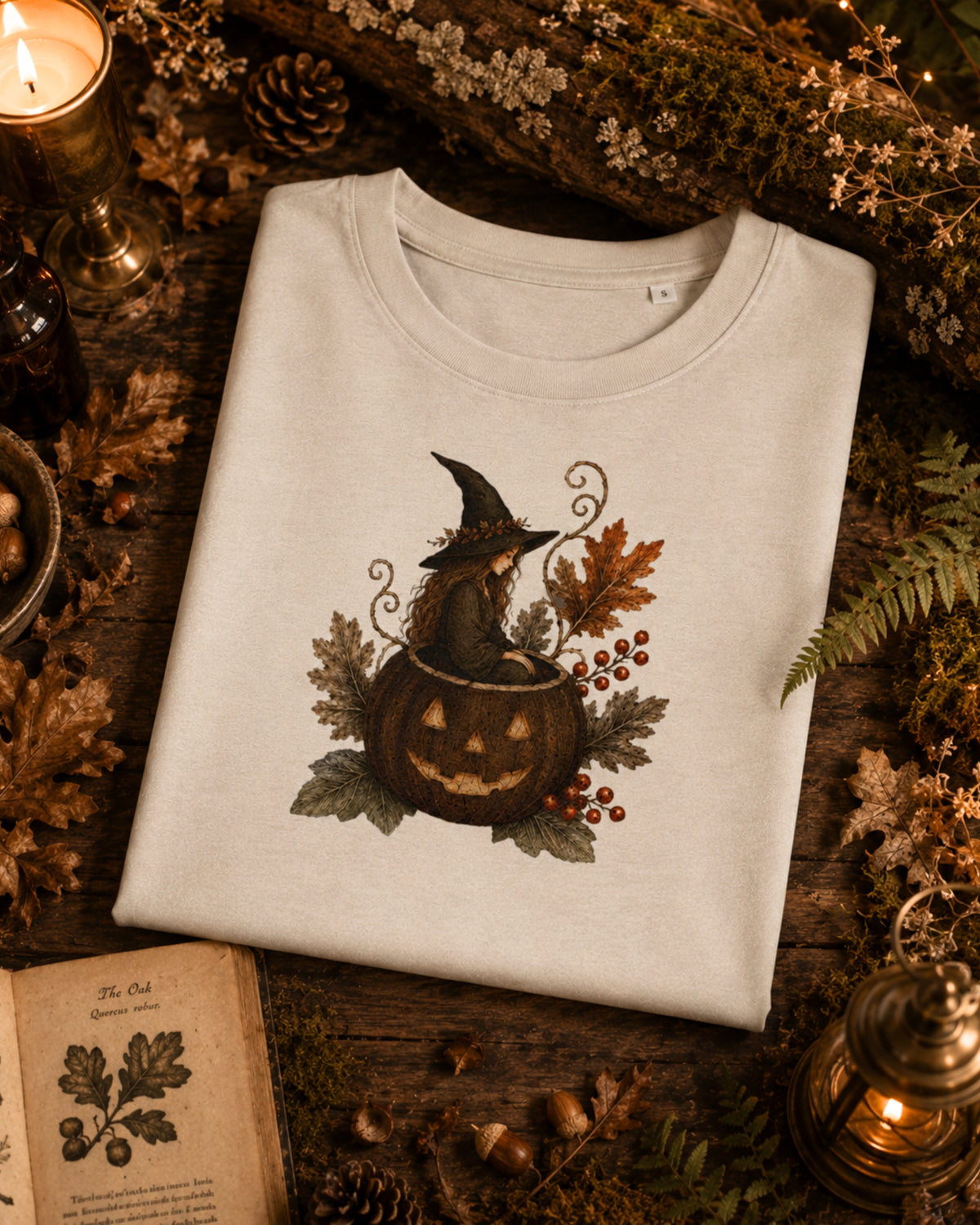 A folded off-white crewneck sweatshirt with a Halloween graphic of a witch sitting in a jack-o'-lantern, surrounded by autumn leaves and berries. The sweatshirt is styled in a cozy, rustic flat-lay setting with a candle, an open book, pinecones, and dried foliage.