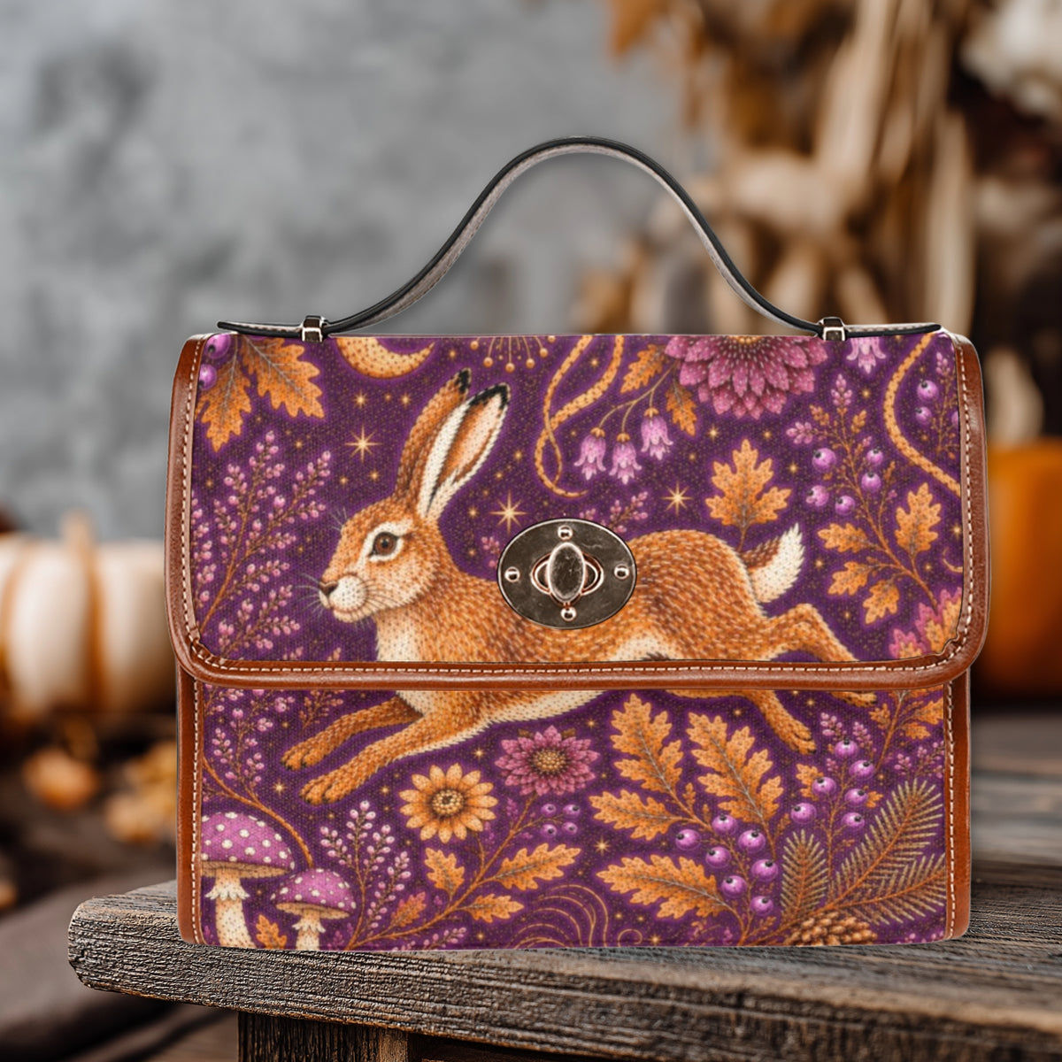 A structured handbag with a top handle, featuring a purple and orange woodland print with a leaping rabbit, flowers, leaves, and mushrooms. The bag has brown leather trim and a round metal clasp, displayed on a rustic wooden surface with a blurred autumn-themed background.