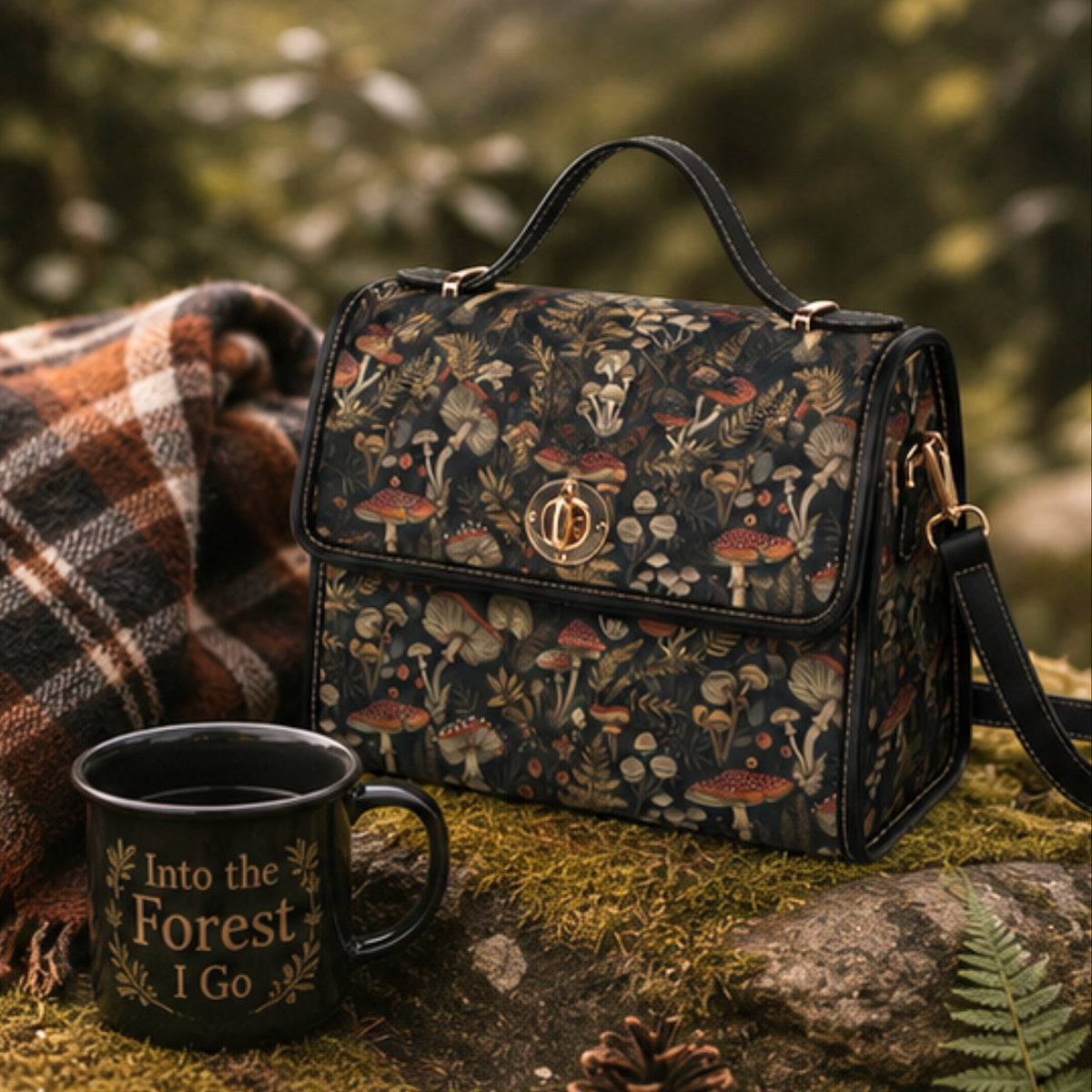 A black handbag with a detailed woodland mushroom print, styled outdoors on mossy rocks next to a black enamel mug that reads 'Into the Forest I Go' and a plaid blanket.