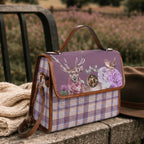 A purple and lavender plaid handbag with a printed front flap featuring a deer, flowers, and a bird, styled outdoors on a stone ledge next to a knit sweater and a brown hat.
