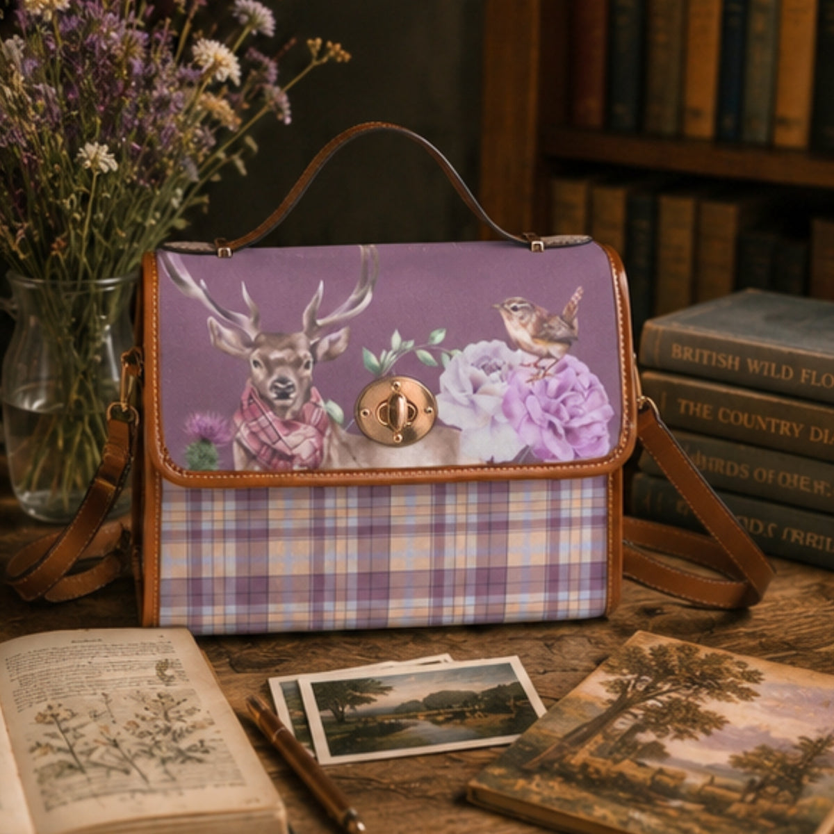 A purple and lavender plaid handbag with a floral and deer print on the front flap, styled on a wooden table with books, dried flowers, and vintage cards.