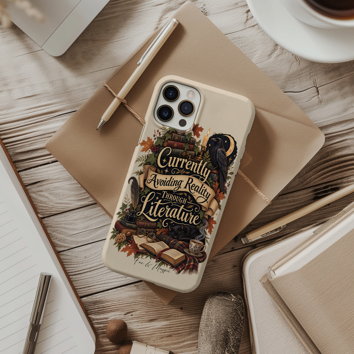 A phone case with a vintage-style bookish illustration and the phrase 'Currently Avoiding Reality Through Literature' is shown in a styled flat-lay on a wooden desk with notebooks, a pen, and a cup of coffee.