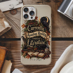 A phone case featuring a vintage-style illustration of a raven, books, and the quote 'Currently Avoiding Reality Through Literature' in a cozy autumn theme, displayed on a rustic wooden table surrounded by props like a camera, hat, and old papers.