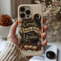 A hand wearing a beige knit sweater holds a cream-colored phone case decorated with a dark raven, books, and the phrase 'Currently Avoiding Reality Through Literature' in a vintage typewriter font. The background is a softly blurred cozy room setting with books and dried flowers.