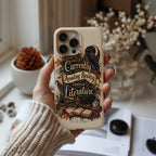A hand wearing a beige knit sweater holds a cream-colored phone case decorated with a dark raven, books, and the phrase 'Currently Avoiding Reality Through Literature' in a vintage typewriter font. The background is a softly blurred cozy room setting with books and dried flowers.