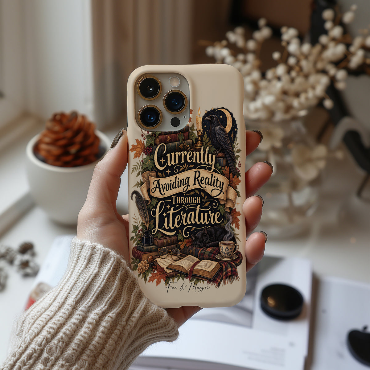 A hand wearing a beige knit sweater holds a cream-colored phone case decorated with a dark raven, books, and the phrase 'Currently Avoiding Reality Through Literature' in a vintage typewriter font. The background is a softly blurred cozy room setting with books and dried flowers.