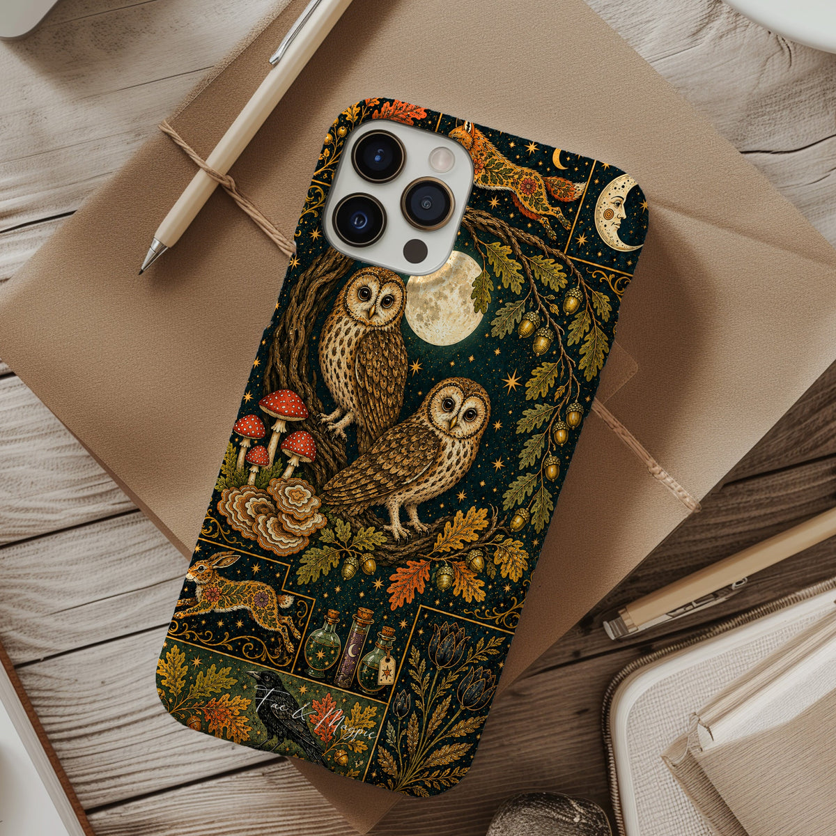 A phone case with a detailed woodland owl illustration pattern, shown in a styled flat-lay on a rustic wooden desk surrounded by notebooks, a pen, and a cup of coffee.