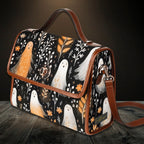 A structured shoulder bag with a Halloween-themed print featuring white ghosts, orange birds, and autumn leaves on a black background, with brown leather trim and handle, displayed on a dark wooden surface.